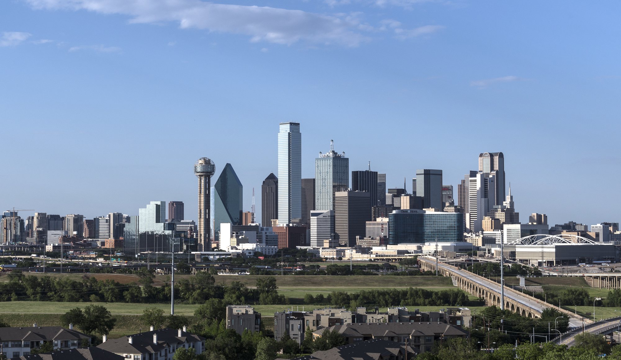 The Dallas skyline under a wide blue sky, seen from south of the Trinity River.